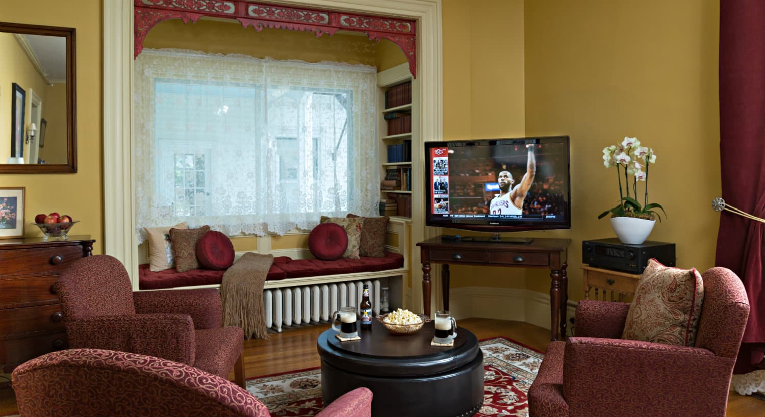 A TV in the corner of a parlor with rose colored chairs surrounded for viewing.