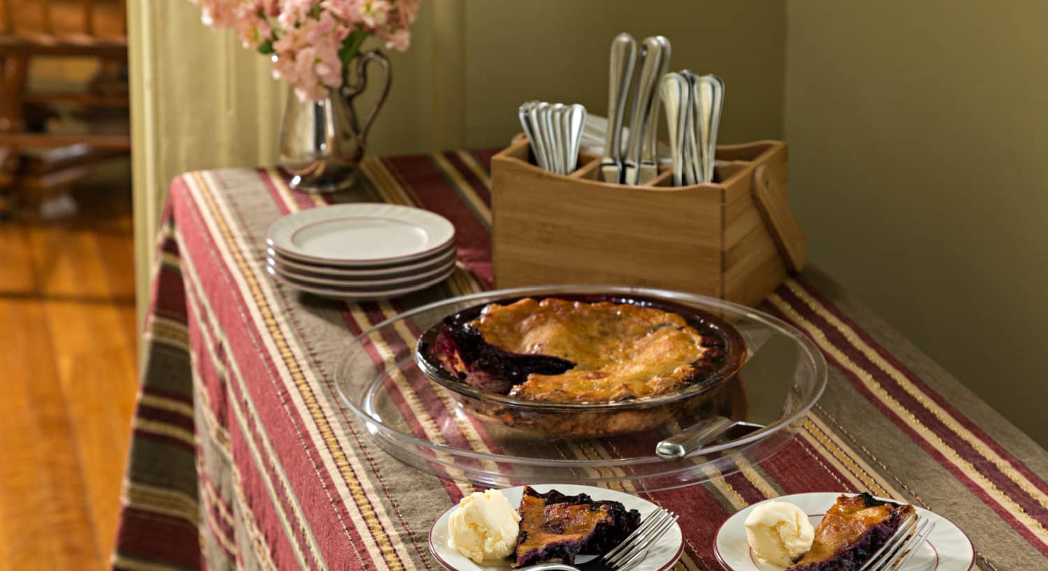 A table with a striped cloth holds a pie along with white plates and cutlery