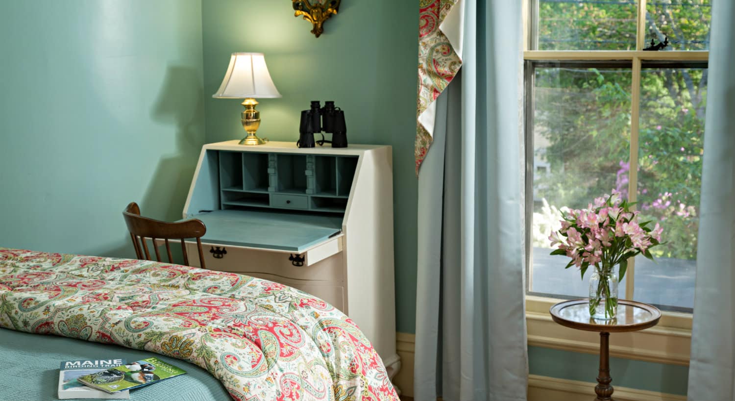 Guest room with pale teal walls holds a bed with a floral throw and a white secretary next to a large window.
