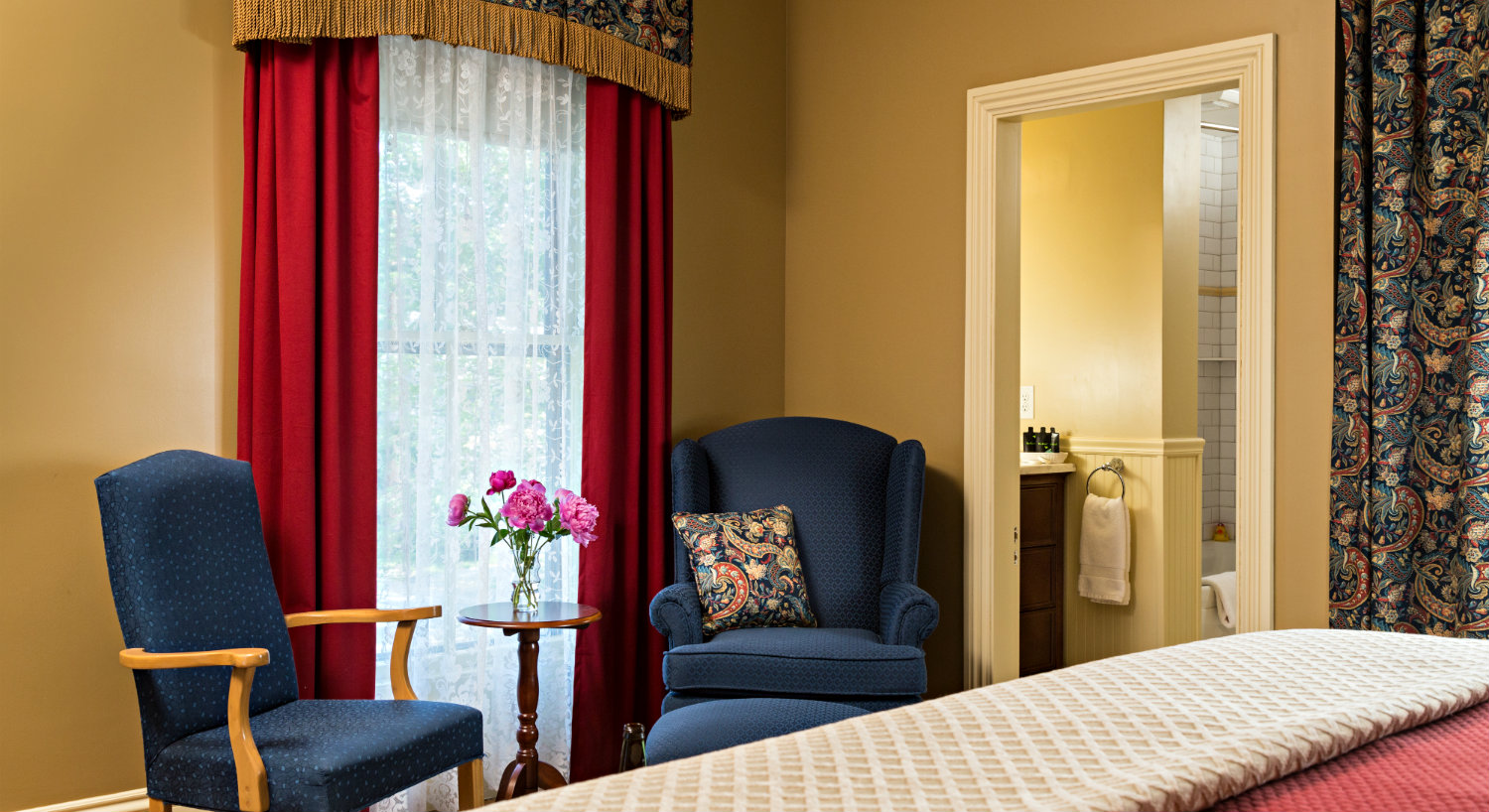 Elegant guest room with beige walls, red curtains and a large bed alongside two blue chairs.