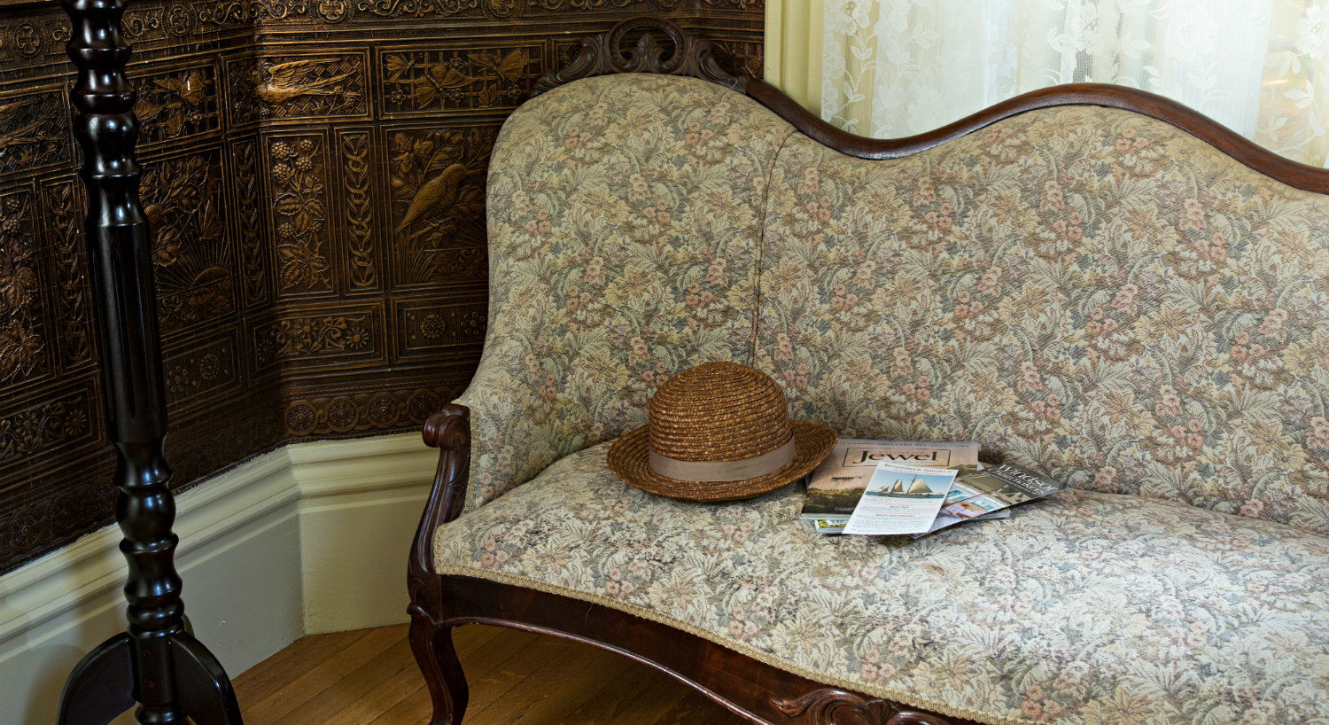 Corner detail of a parlor area with an antique sofa in cream and a window with lace curtains.