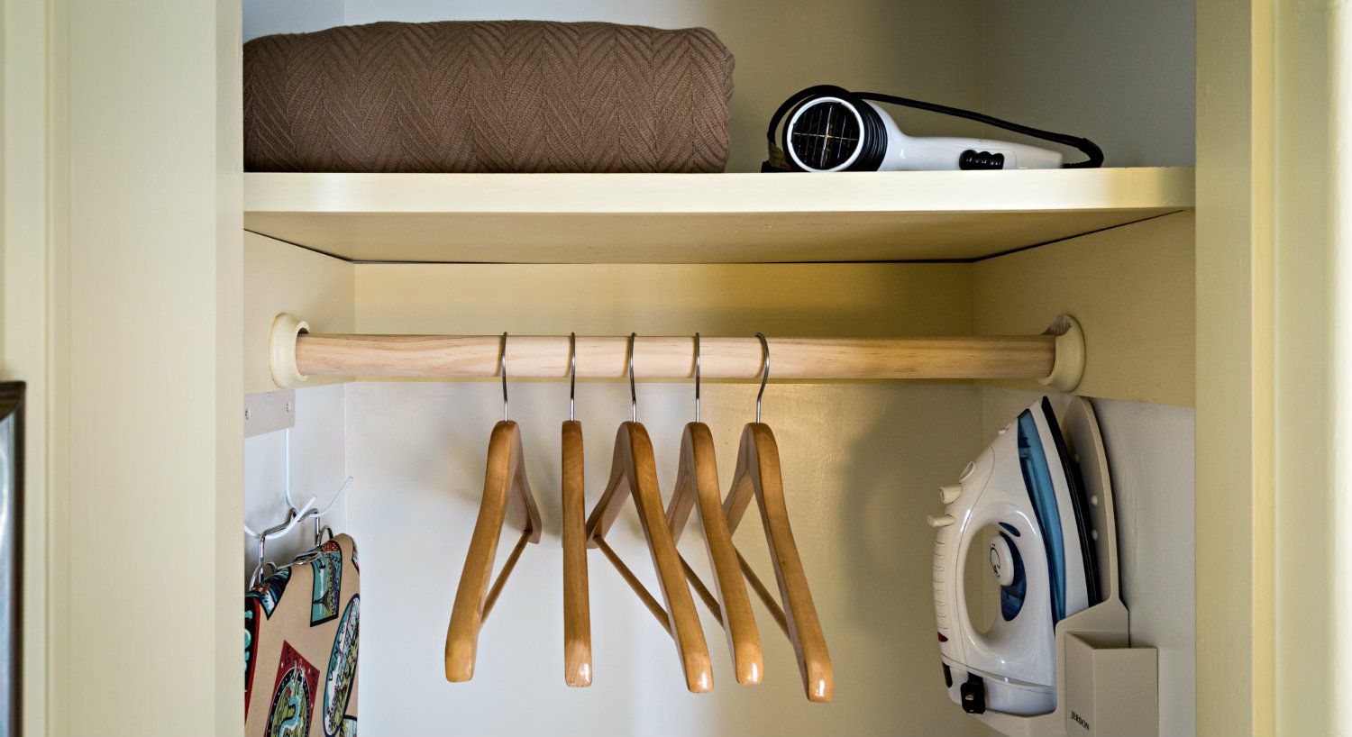 An open closet which holds wooden hangers, and iron and ironing board and extra blankets.