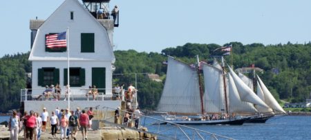 People walk along the pier near a lighthouse with sailboats in the background.