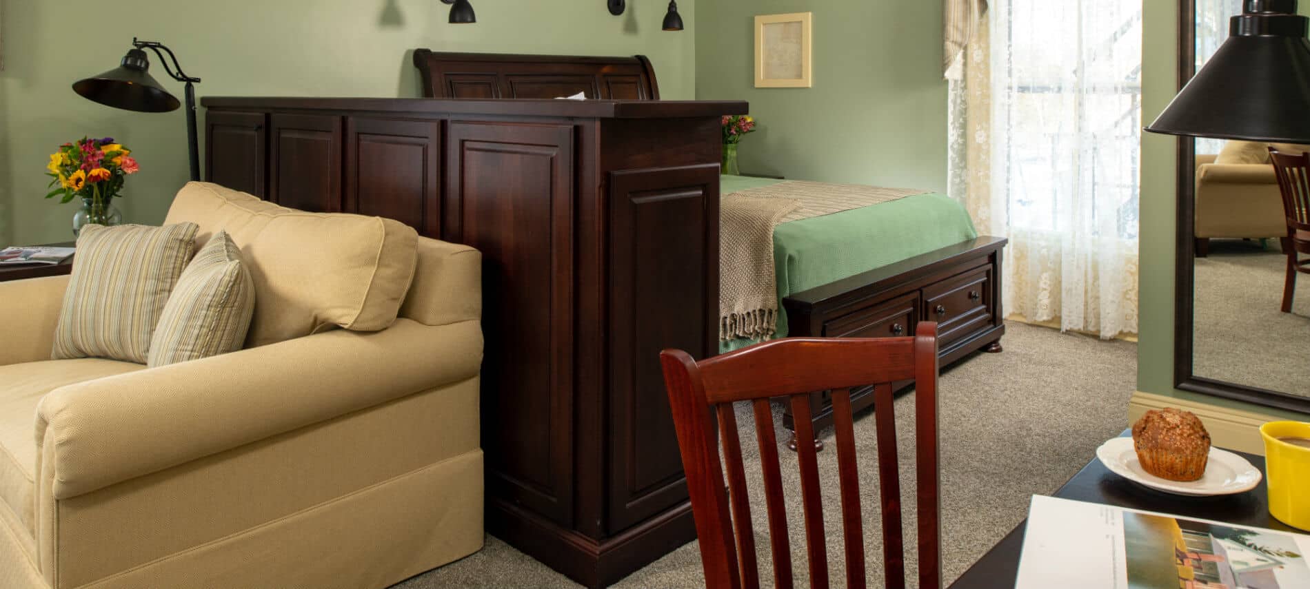 Guestroom with green walls has a seating and desk area separated from the bed with a large cherry wood cabinet.