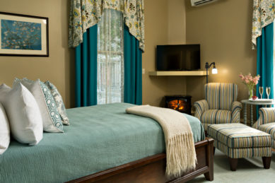 A room with beige walls and soothing blue and green accents on the bedding and chair fabric with a small fireplace and TV.
