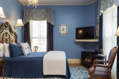 A blue bedroom with an antique wooden bed, a TV, desk, side chair and a small iron fireplace.