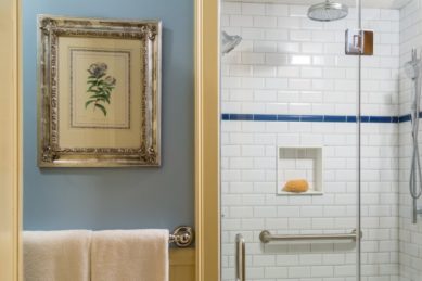A shower tiled in white in a blue bathroom with a painting and towels.