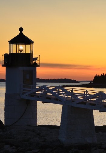 A lit lighthouse looks over the harbor in a hazy orange sunset.