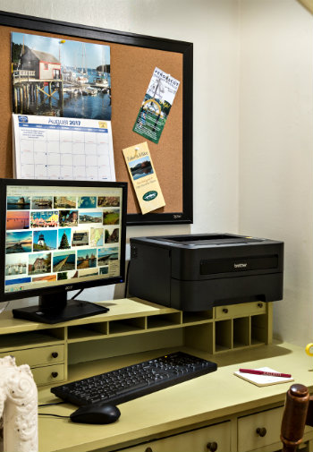 A yellow desk set up with a desktop monitor and keyboard as well as a printer with a calendar on a corkboard behind.