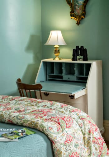 A cozy bed next to a cream colored secretary desk in a room with pale green walls.