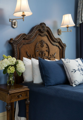 Ornate wooden headboard on a bed made up in blue bedding with several pillows in a room with blue walls and light sconces.
