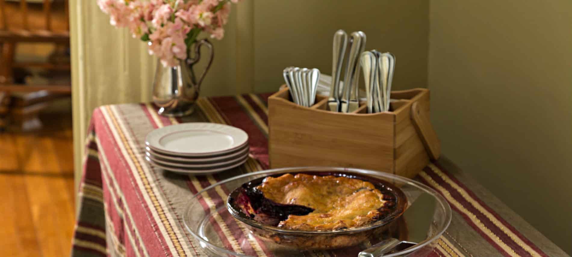 A table with a striped cloth holds a pie along with white plates and cutlery. .